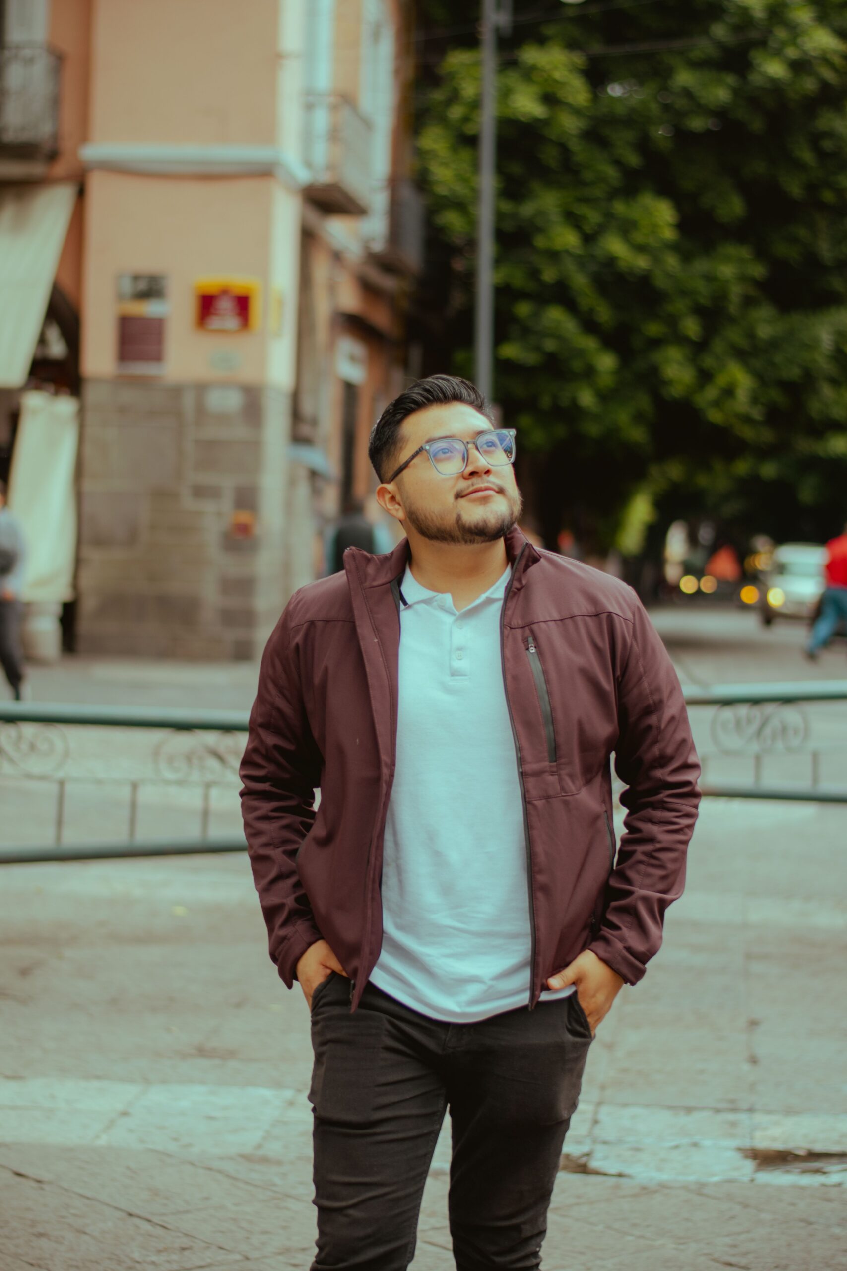 A stylish young man in a city street wearing glasses and a jacket, looking thoughtful.